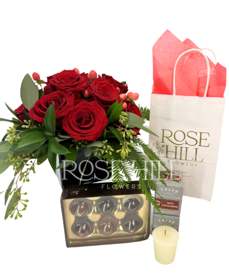 Cluster of red roses in a cube vase, votive candles by TRAPP Fragrances, and a box of truffles.