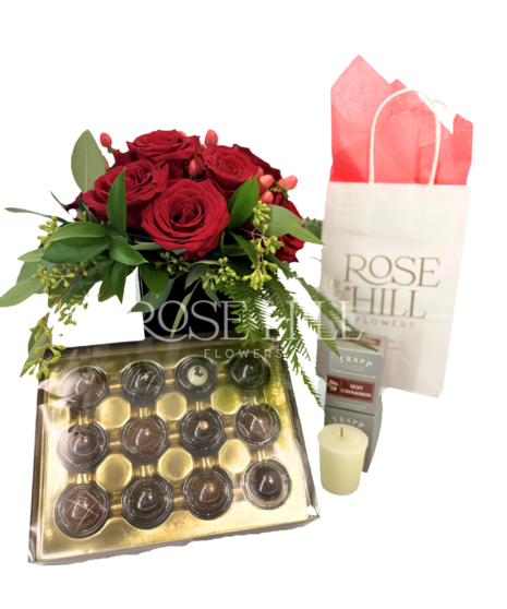 Cluster of red roses in a cube vase, votive candles by TRAPP Fragrances, and a box of truffles.