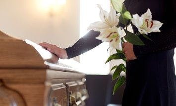 A person in black clothing holds white lilies, placing a hand on a wooden coffin inside a softly lit room with light-colored walls.